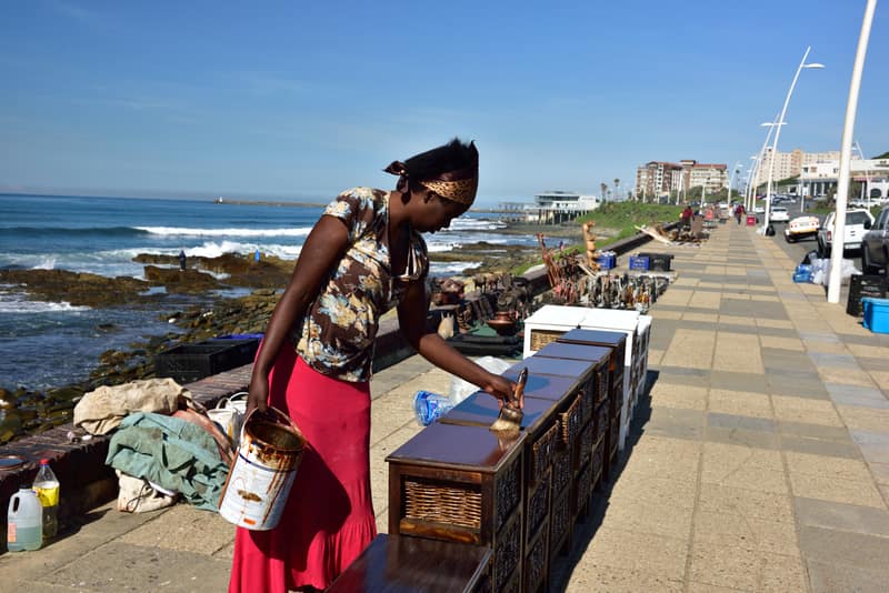 A trader setting out her stall on the East London beachfront promenade, the sea behind her.