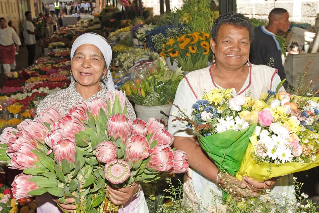 Two flower sellers in Cape Town smiling over armfuls of king proteas and mixed bouquets at their stall.